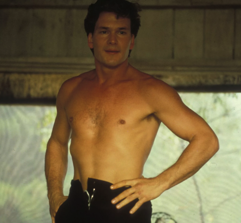 Patrick Swayze as Johnny Castle, a light-skinned man with short, styled brown hair, is standing topless, wearing a pair of unzipped, black pants with his hands on his hips.
